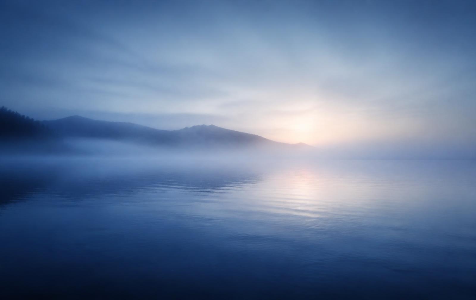 Still blue water at dawn with soft mist over distant hills